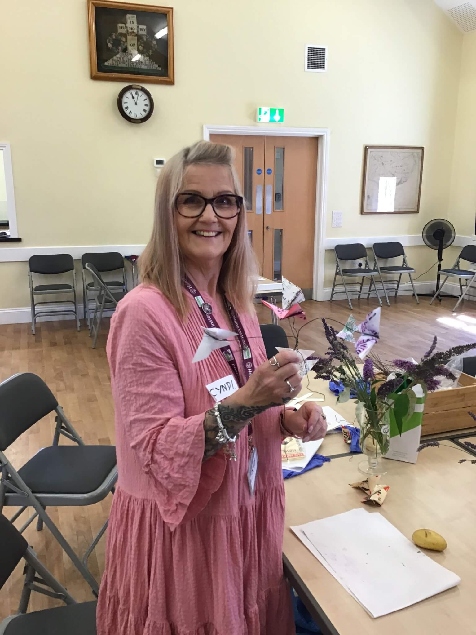 A woman in a pink dress standing in a room with chairs and tables, holding a small item, smiling at the camera. - Home Instead