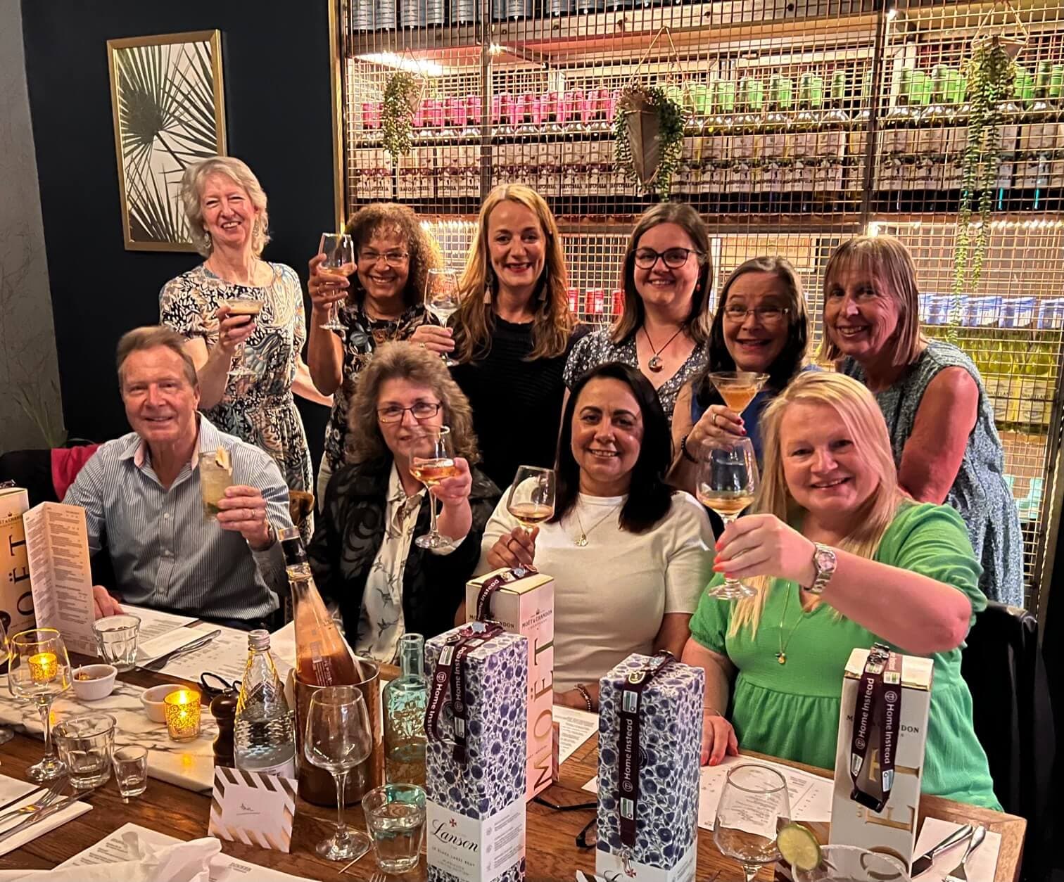 A group of people raise their glasses and smile while seated at a table filled with gifts and drinks in a cozy restaurant. - Home Instead