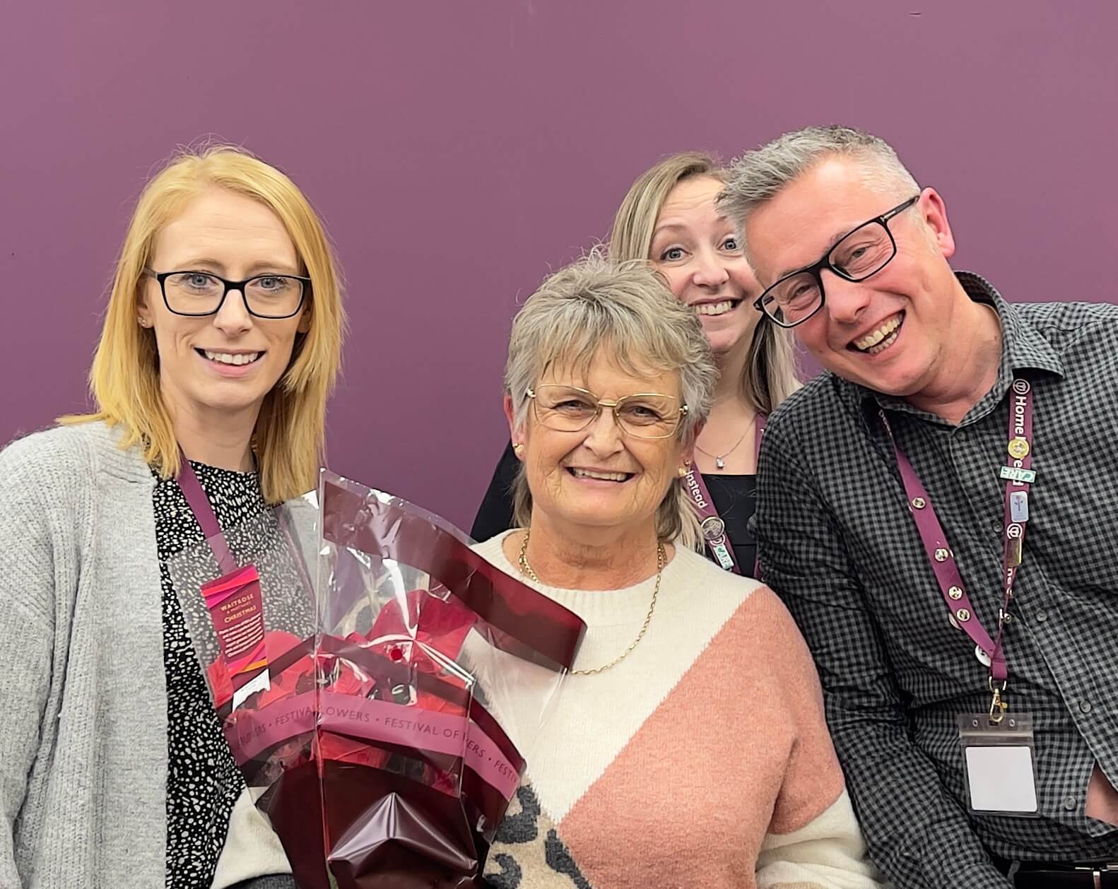 Four people smiling in front of a purple wall; woman in center holding floral wrapped gift. - Home Instead