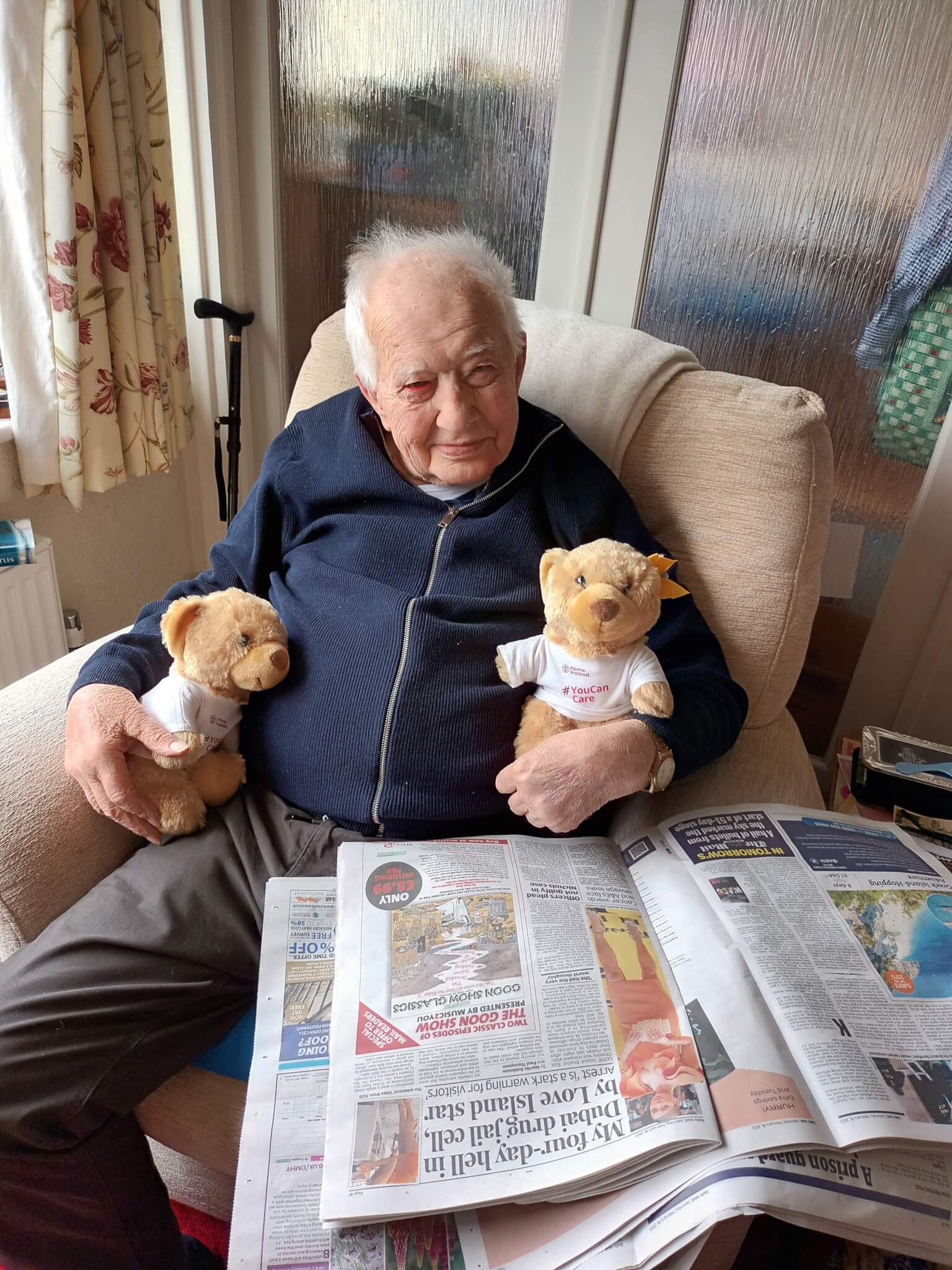 An elderly man sits in a chair, holding two teddy bears. A newspaper is open on his lap. - Home Instead