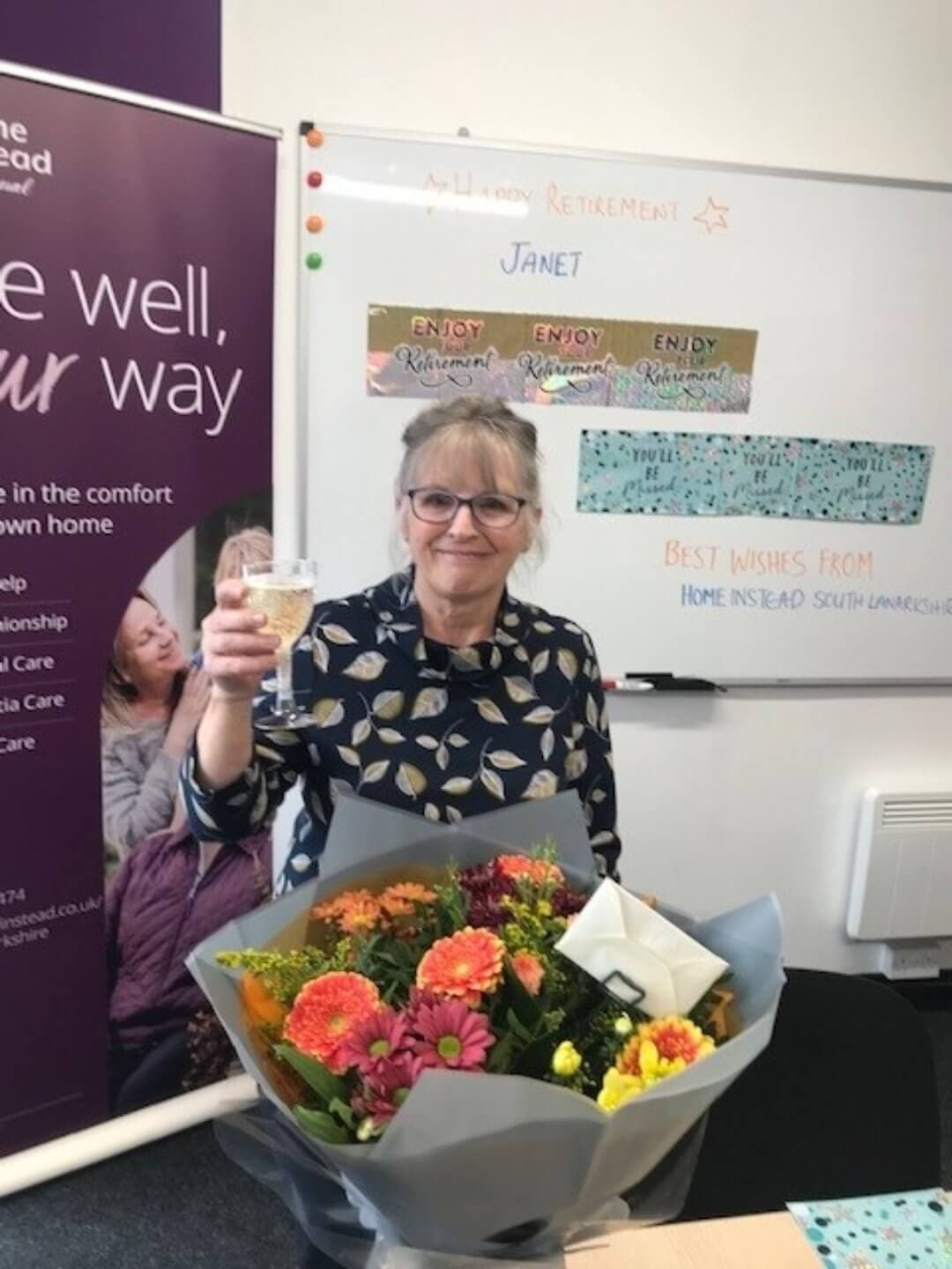 Woman holding a glass and flowers, celebrating retirement in an office with congratulatory signs and messages. - Home Instead