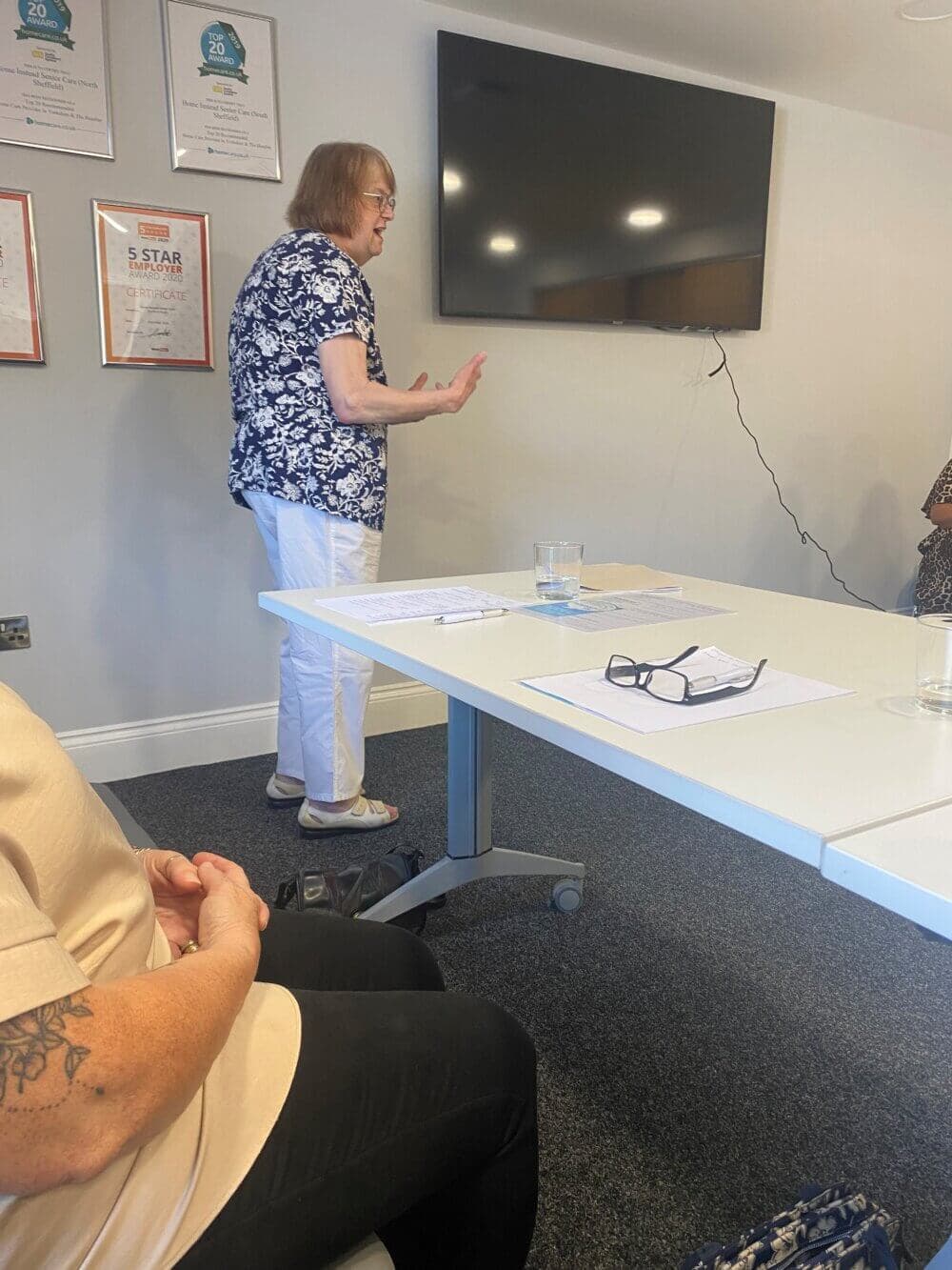 Person in a floral shirt giving a presentation by a screen, with others seated at a table in an office setting. - Home Instead