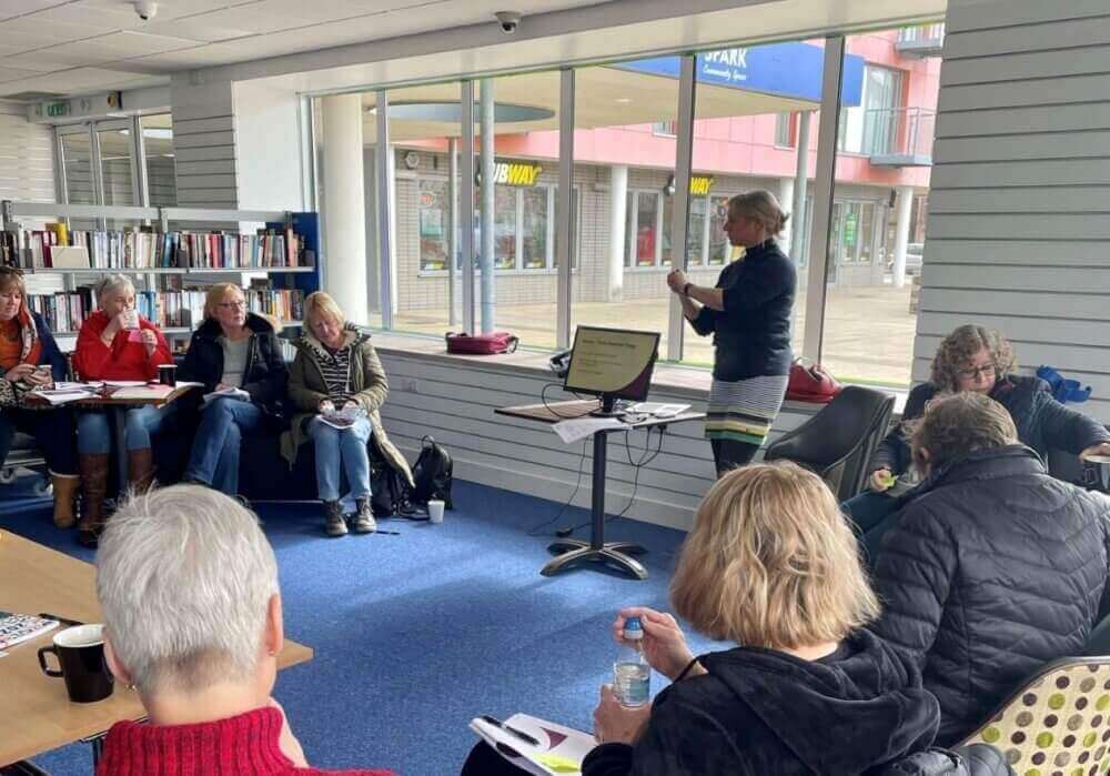 A woman stands at a presentation while others are seated in a library, listening attentively and taking notes. - Home Instead