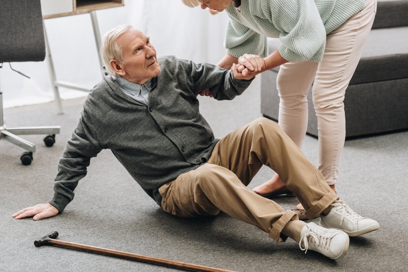 Elderly man sitting on the floor with a cane nearby, being helped up by a woman. - Home Instead