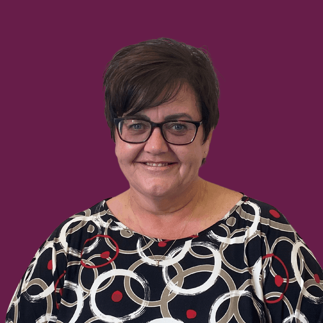 A woman with short hair and glasses wearing a patterned shirt smiles in front of a purple background. - Home Instead