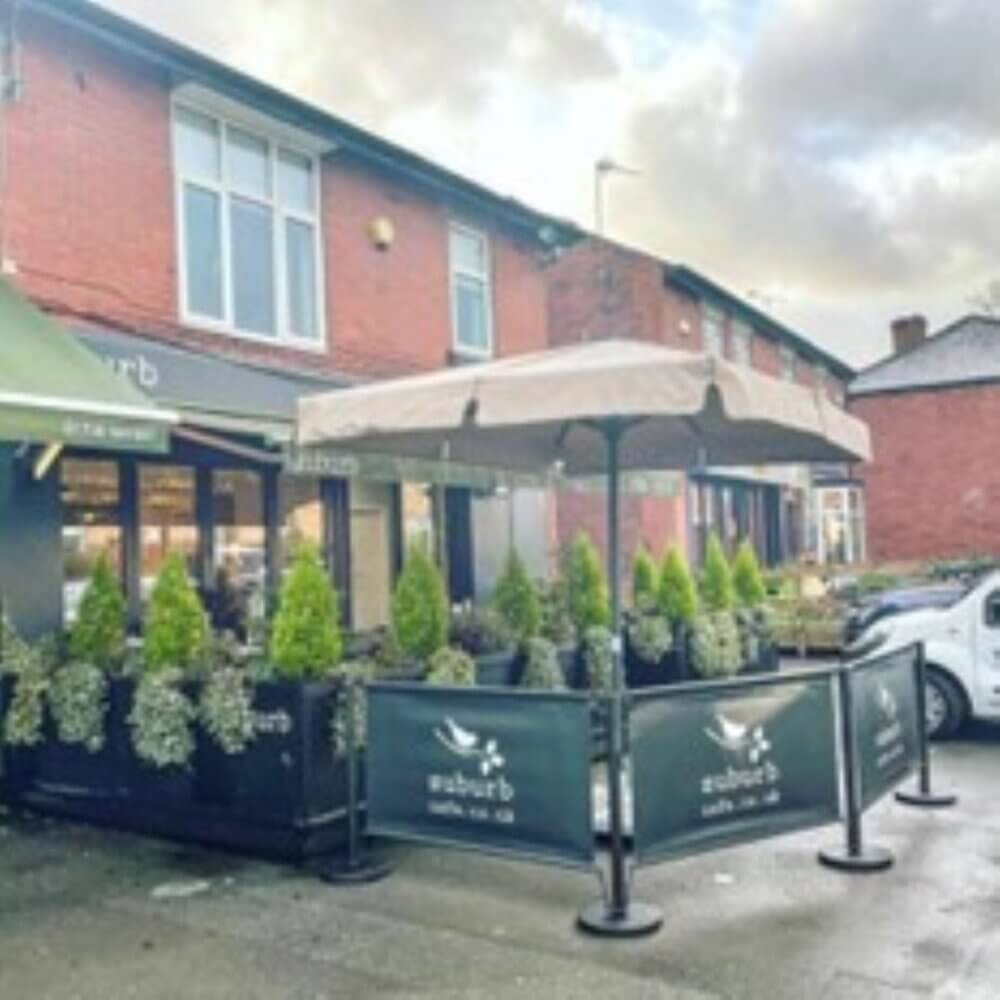 A small cafe with outdoor seating, potted plants, and an umbrella, situated in front of a brick building. - Home Instead