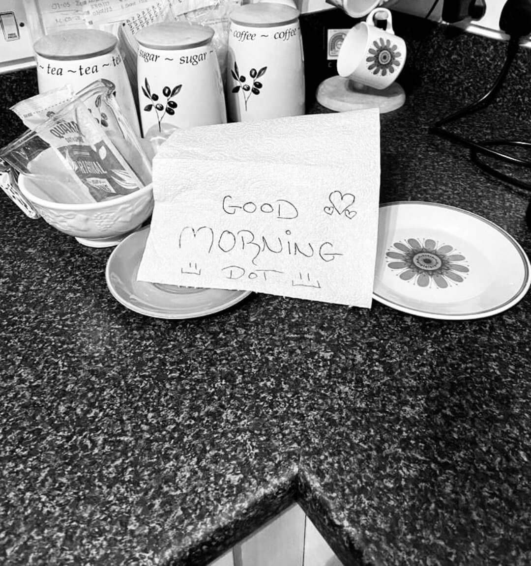 A handwritten note reading "Good Morning Dad" is placed on a kitchen counter with cups and jars in the background. - Home Instead