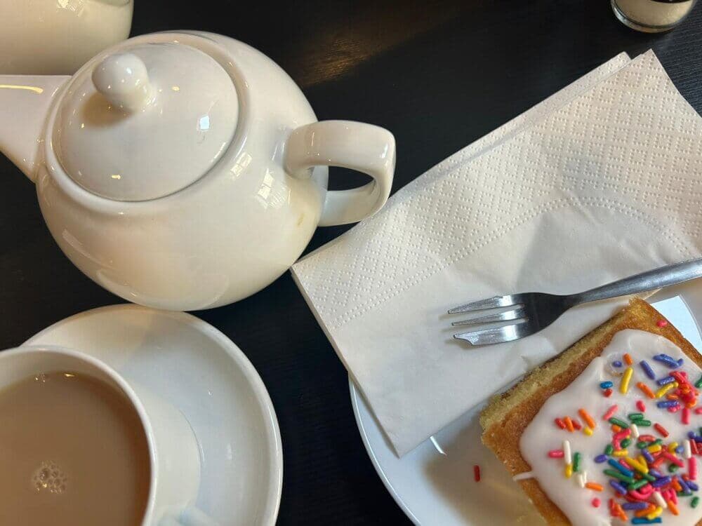 A teapot, teacup with tea, and a slice of cake with white icing and sprinkles on a plate with a fork and napkin. - Home Instead