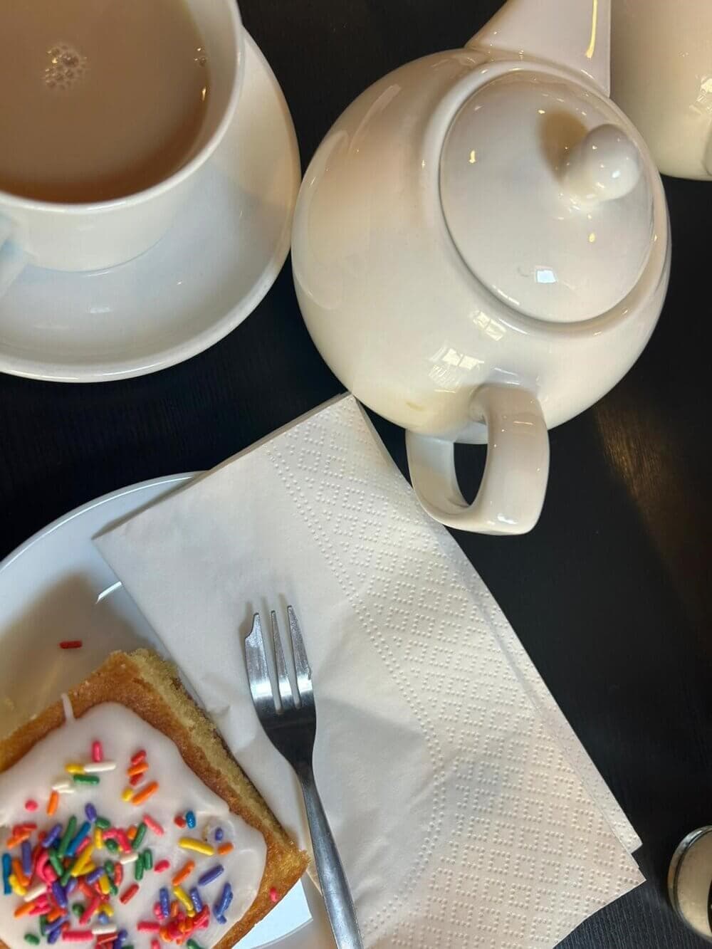 A cup of tea and a teapot next to a fork and a pastry with icing and rainbow sprinkles on a white plate. - Home Instead