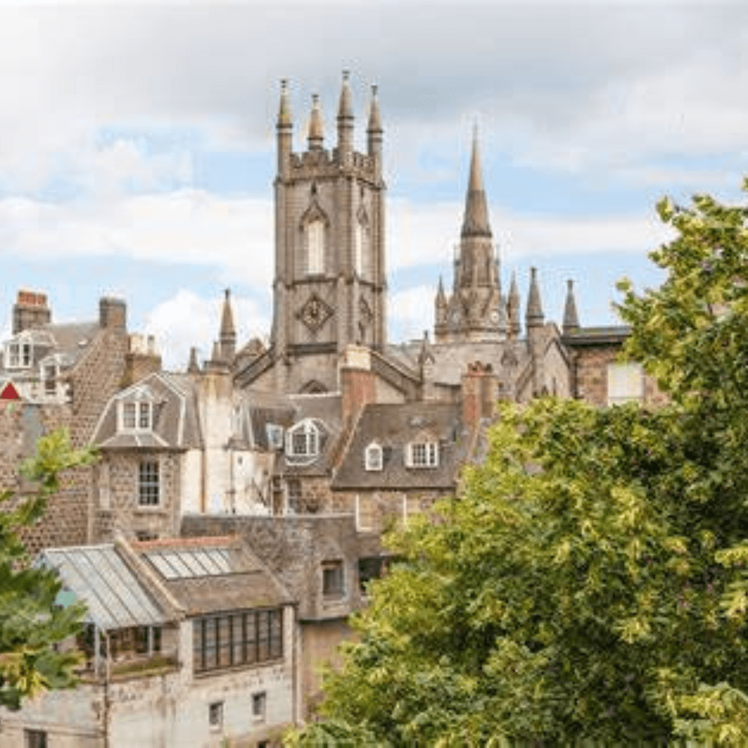 A view of a historic city with prominent church towers, old stone buildings, and lush green trees in the foreground. - Home Instead