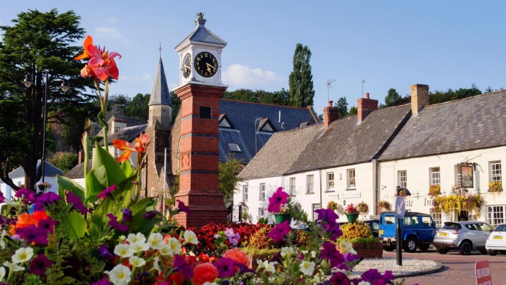 A picturesque village clock tower with vibrant flowers in the foreground and charming houses in the background. - Home Instead