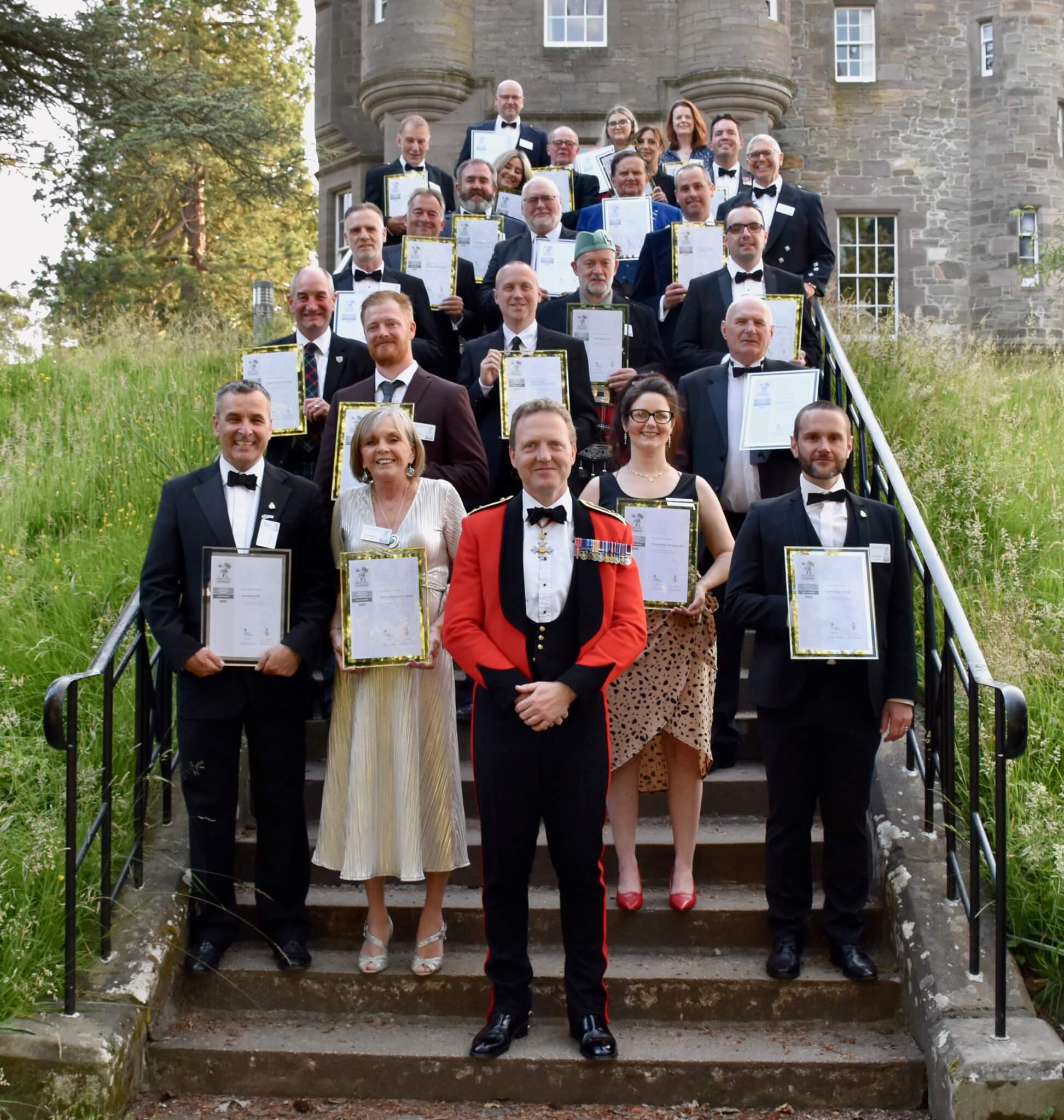 A group of people in formal attire standing on steps, holding certificates in front of a historic stone building. - Home Instead