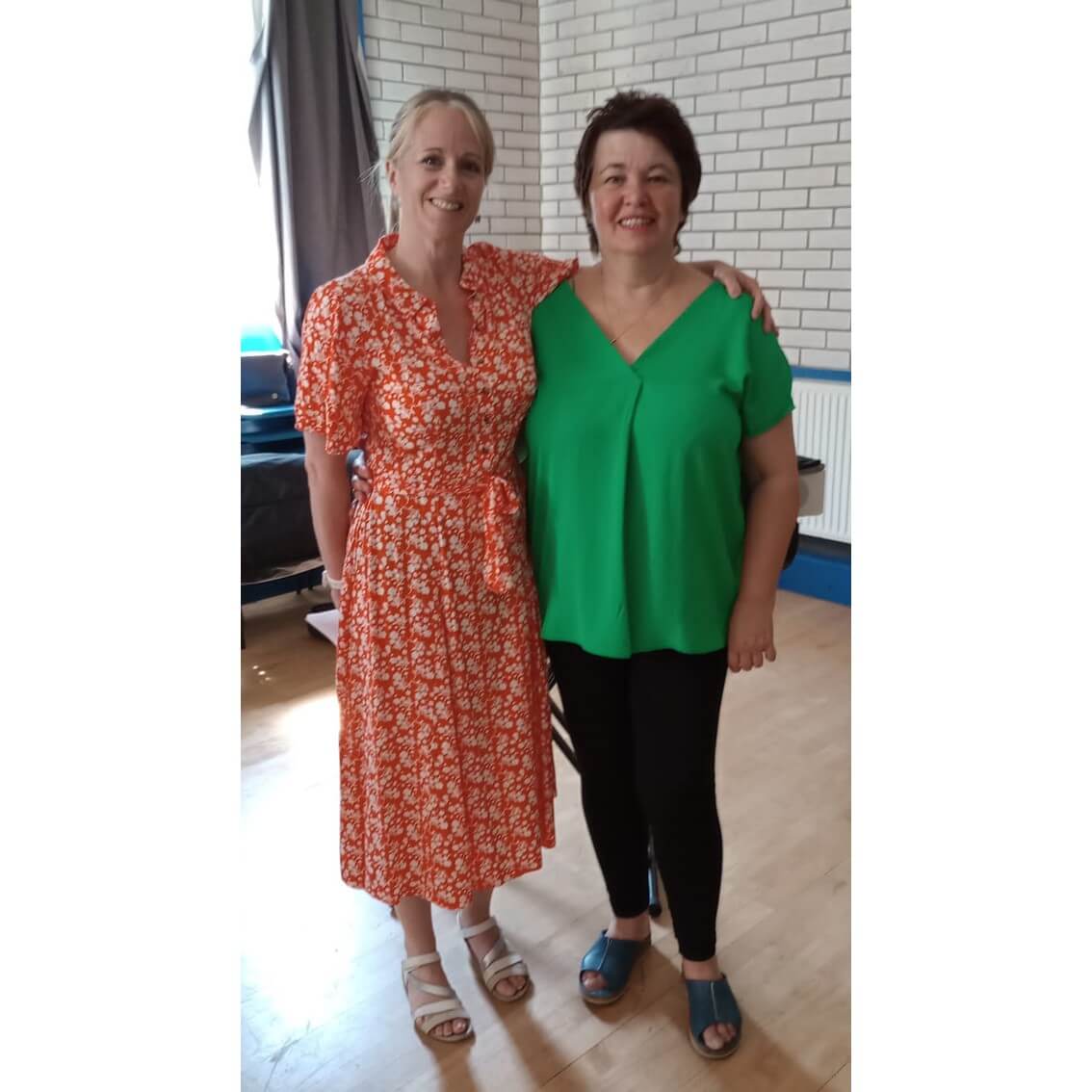 Two women standing close together smiling, one in an orange floral dress and the other in a green top with black pants. - Home Instead