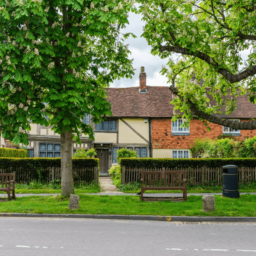 A quaint house with a mix of brick and timber facade, lush green trees, benches, and a paved path in the foreground. - Home Instead
