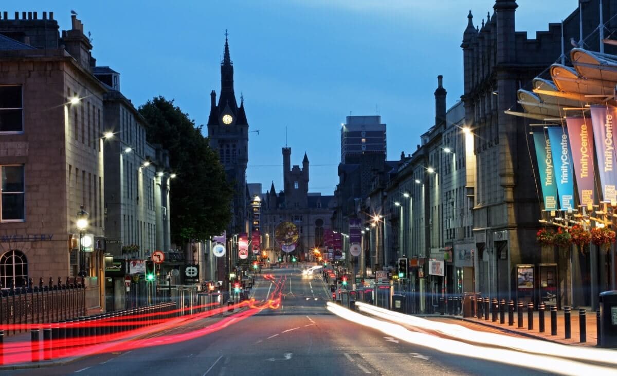 A dimly lit, busy city street at dusk with light trails from cars and historic buildings in the background. - Home Instead
