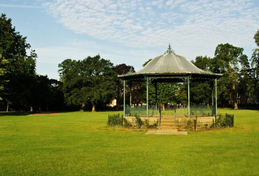 A Victorian-style gazebo in the middle of a green park with trees in the background under a partly cloudy sky. - Home Instead
