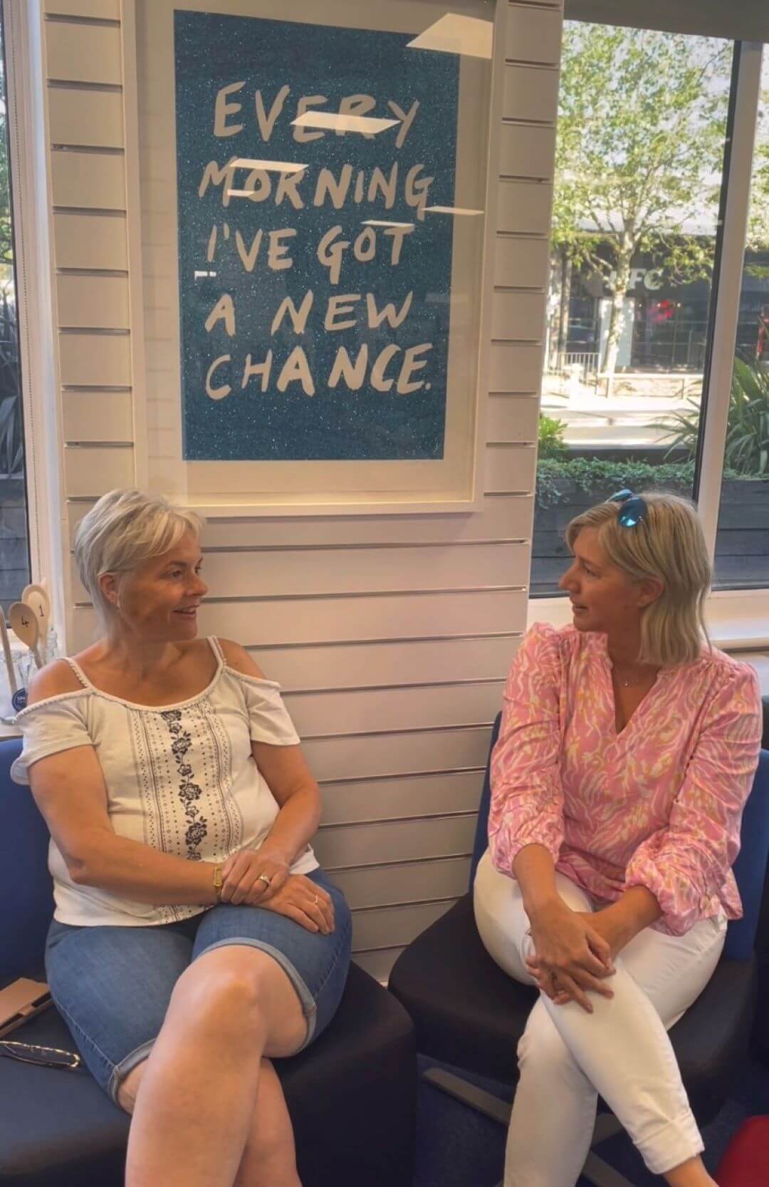 Two women sit on benches in front of a sign that reads "EVERY MORNING I'VE GOT A NEW CHANCE." They are talking and smiling. - Home Instead