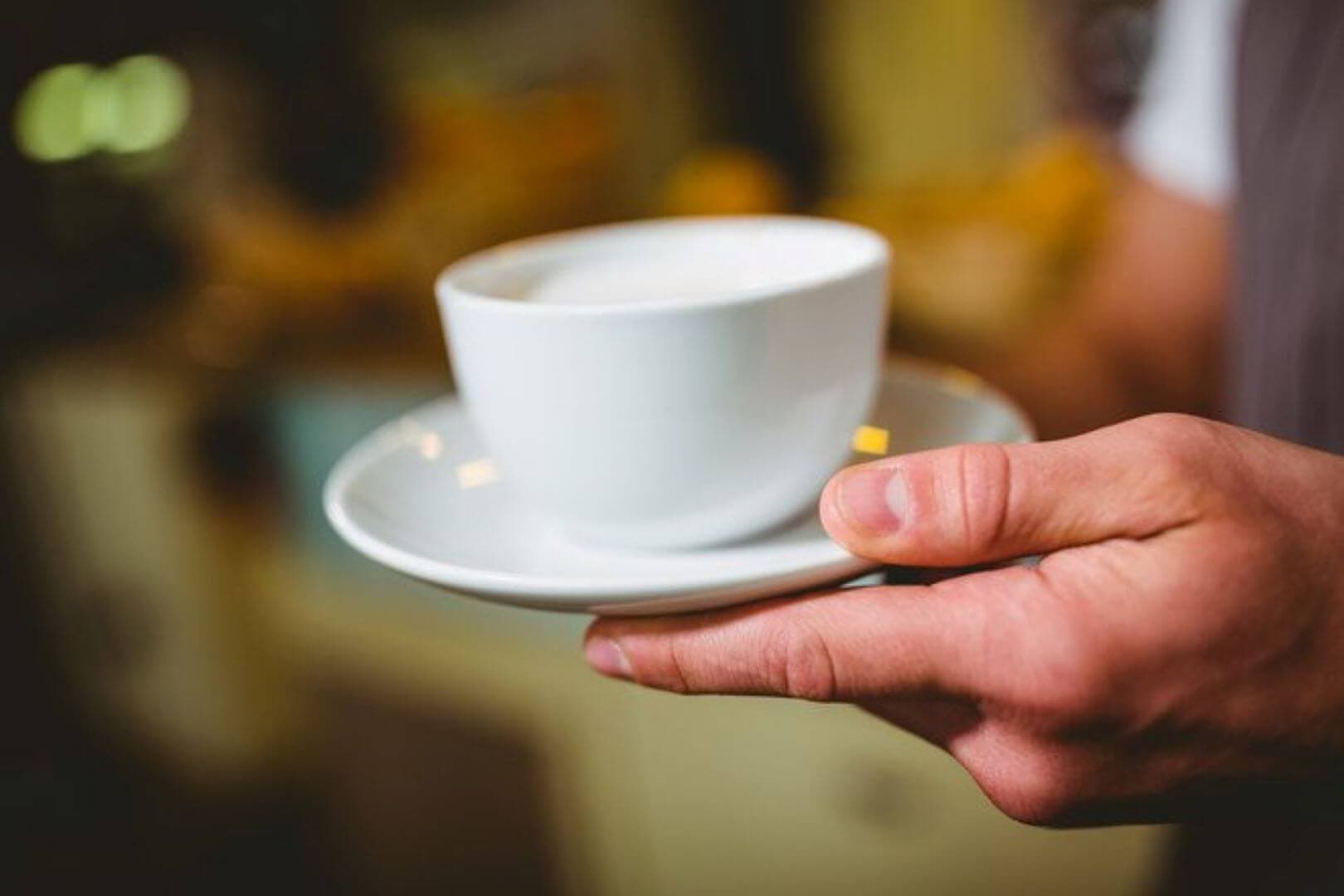 A close-up of a person's hand holding a white cup and saucer with a hot beverage, likely coffee or tea. - Home Instead