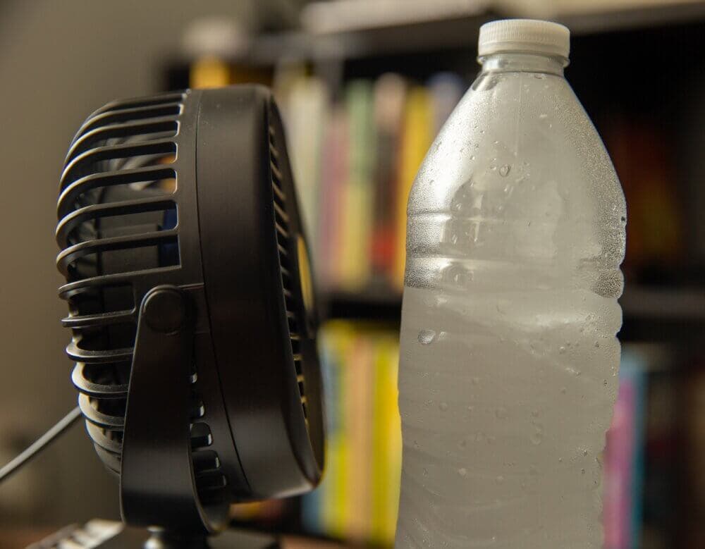 A small fan beside a cold, condensation-covered water bottle with a blurred bookshelf in the background. - Home Instead