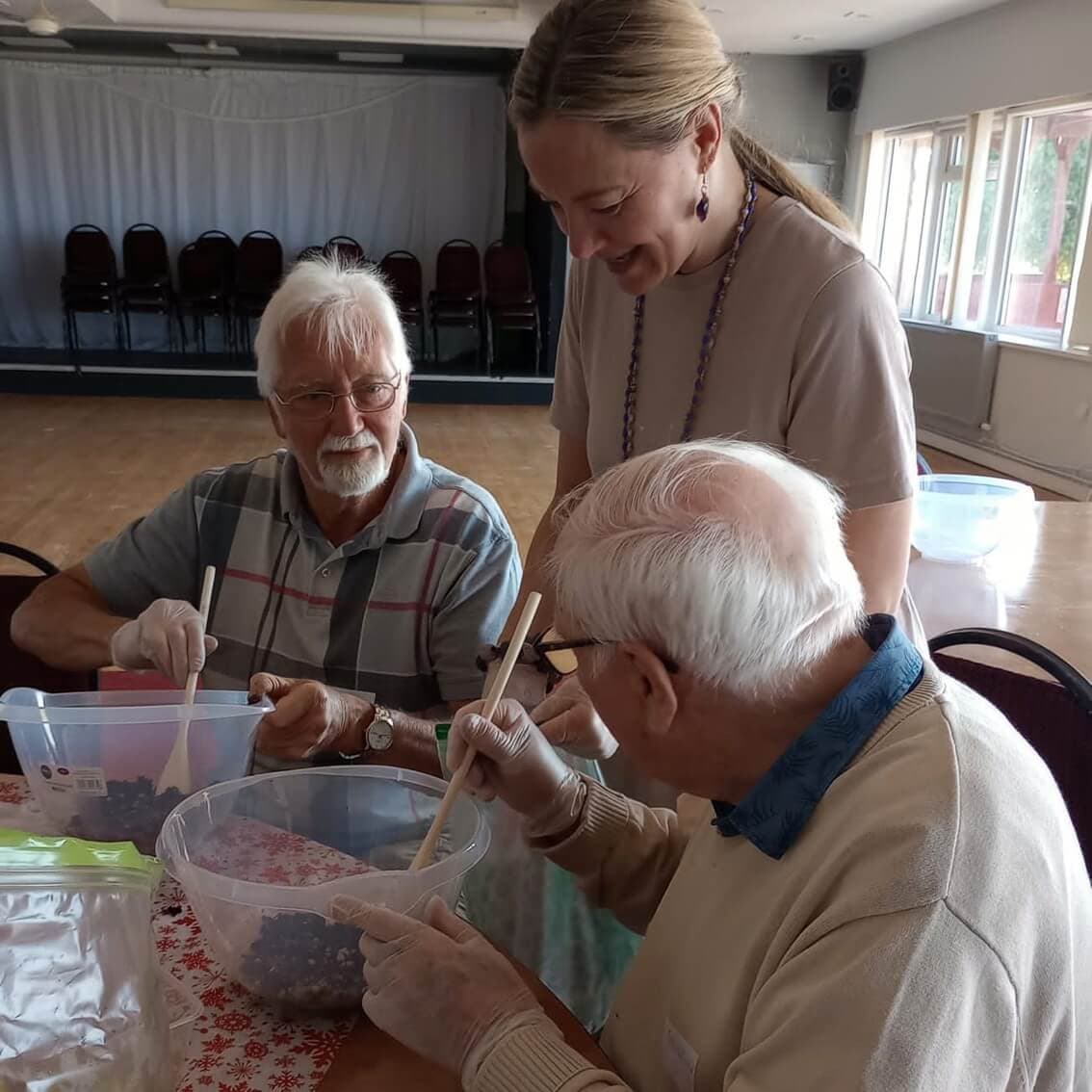 Three elderly people work together in a community center, mixing ingredients in large bowls while wearing gloves. - Home Instead