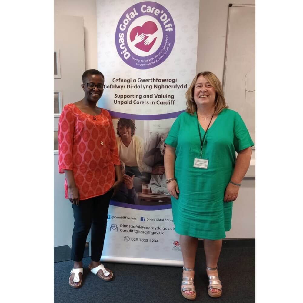 Two women stand smiling in front of a banner for "Dinas Gofal Care Diff" supporting unpaid carers in Cardiff. - Home Instead