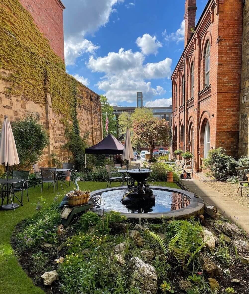 A courtyard garden with a fountain, tables, chairs, and umbrellas, surrounded by brick buildings and greenery under a blue sky. - Home Instead