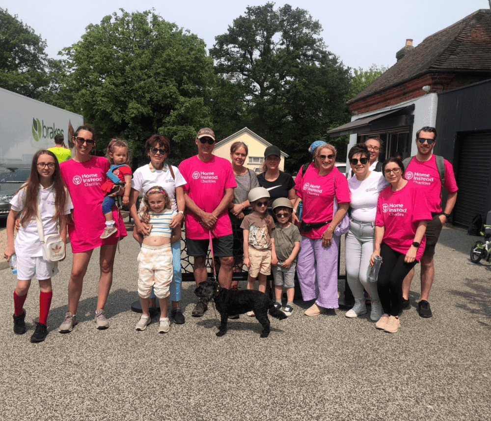 A diverse group of people, including kids and a dog, pose outdoors wearing bright pink shirts. Some wear hats and sunglasses. - Home Instead