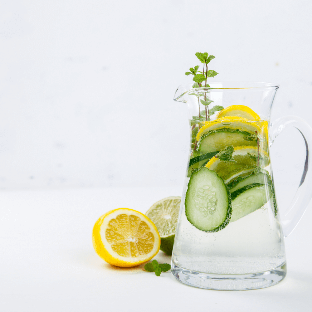 A glass pitcher of water with lemon slices, cucumber slices, and mint leaves, next to halved lemons and limes. - Home Instead