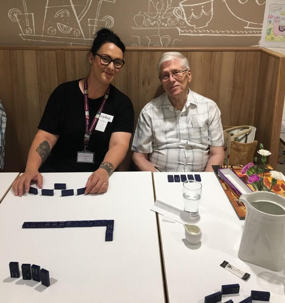 Two people sitting at a table, smiling, and playing dominoes in a cozy room. - Home Instead