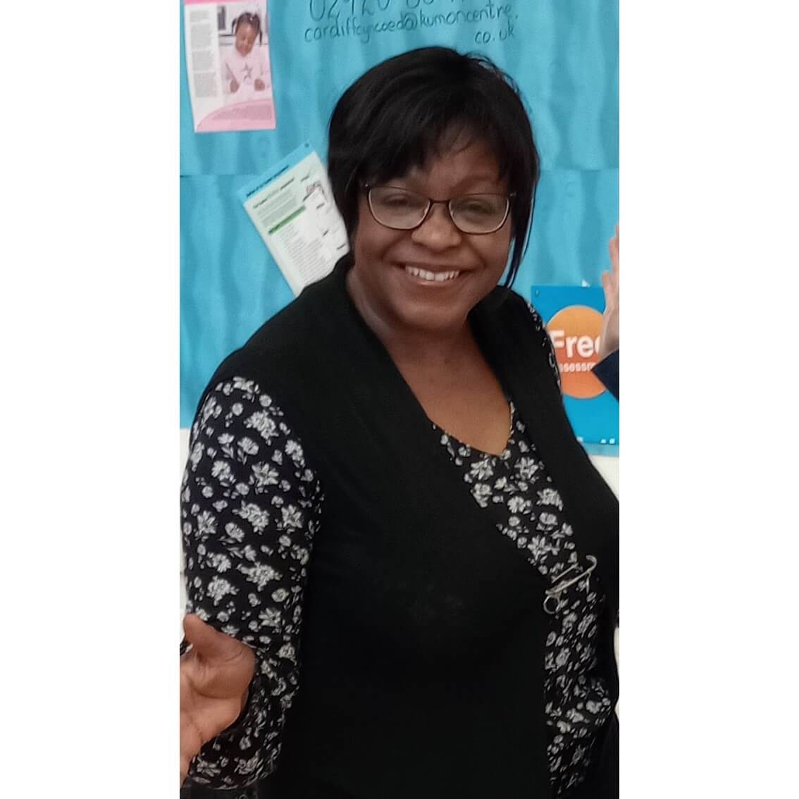 A smiling woman with glasses and a black vest stands in front of a teal background with posters. - Home Instead