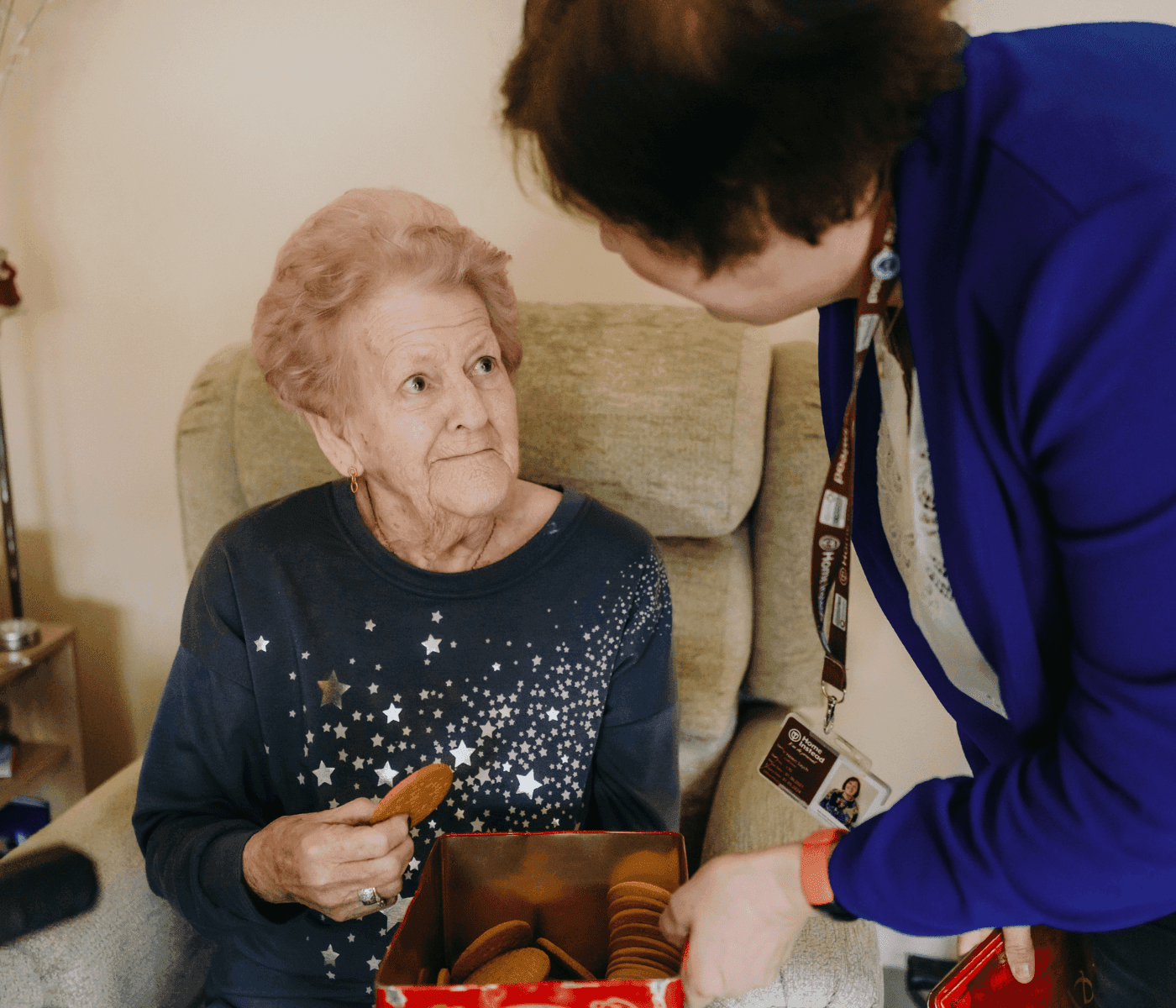 An elderly woman seated, holding a cookie, while a standing woman in blue talks to her. - Home Instead