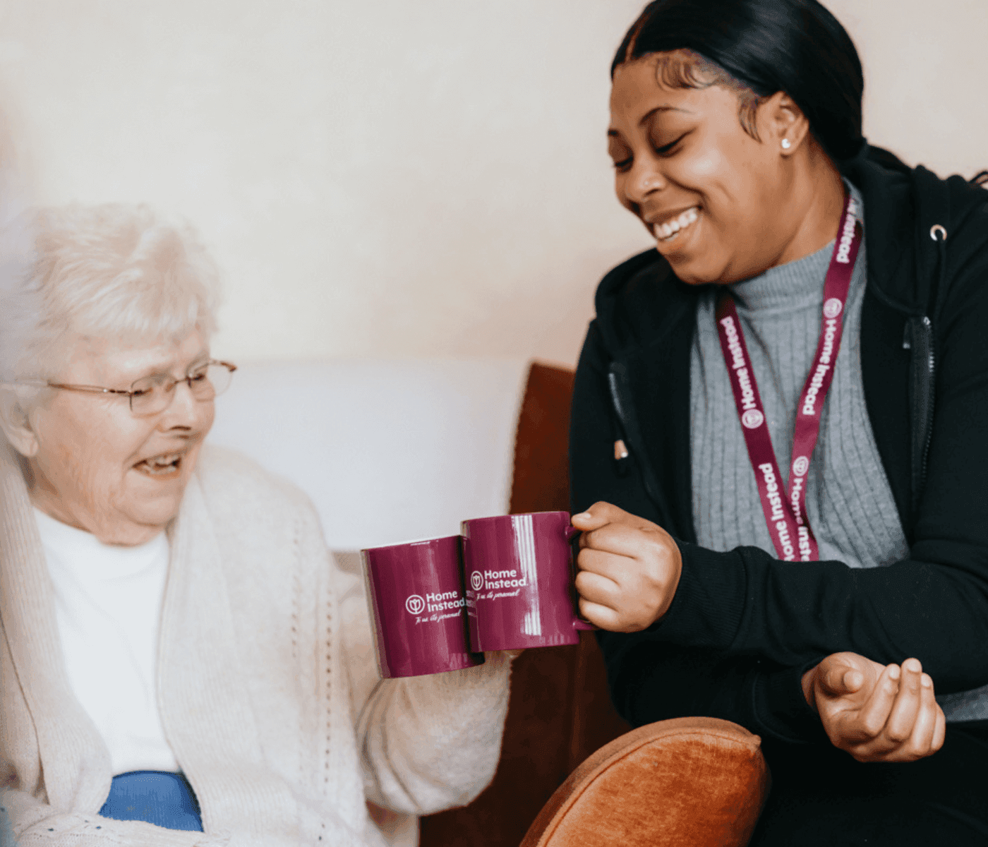 Two women, one elderly and one caregiver, smiling and clinking mugs with "Home Instead" branding while seated. - Home Instead