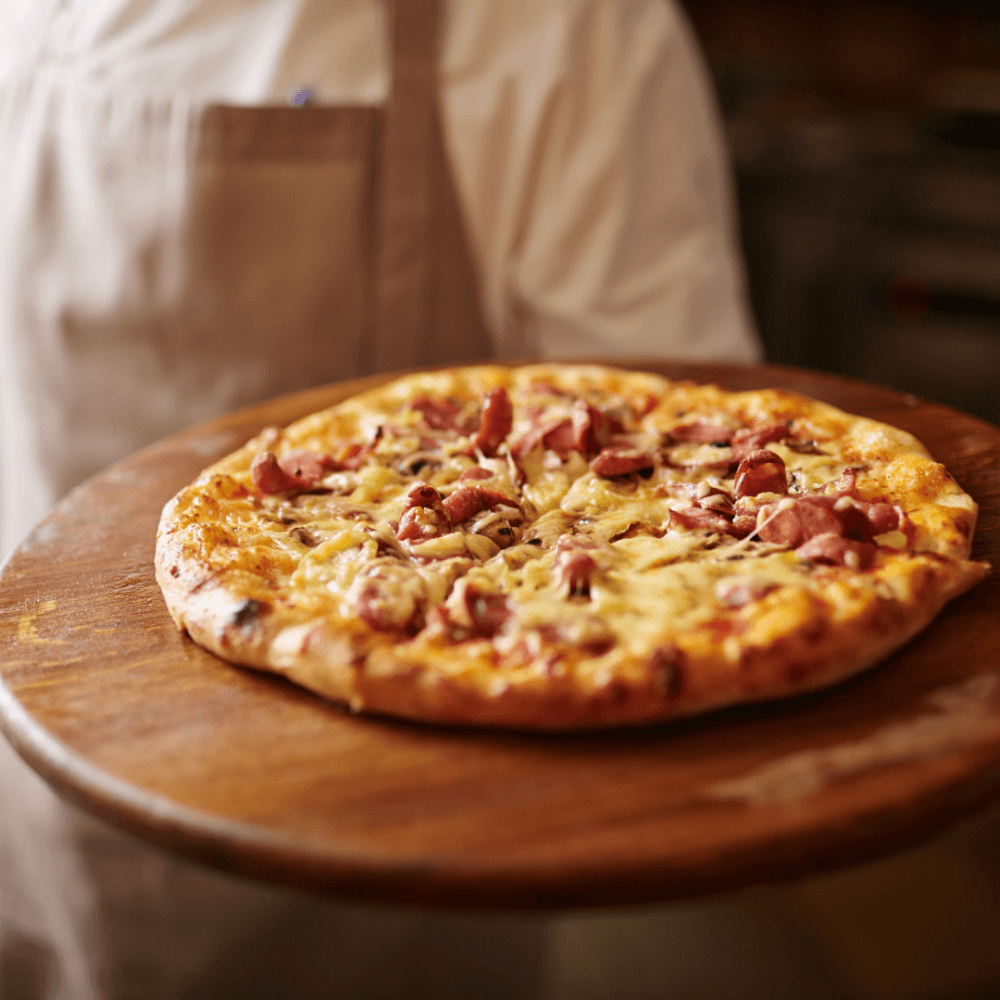 A person holds a freshly baked pizza topped with ham and cheese on a wooden board. - Home Instead