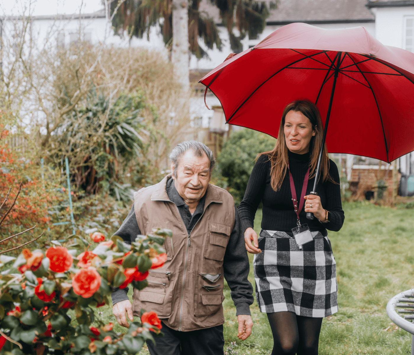 A woman holding a red umbrella walks with an elderly man in a garden with red flowers. - Home Instead
