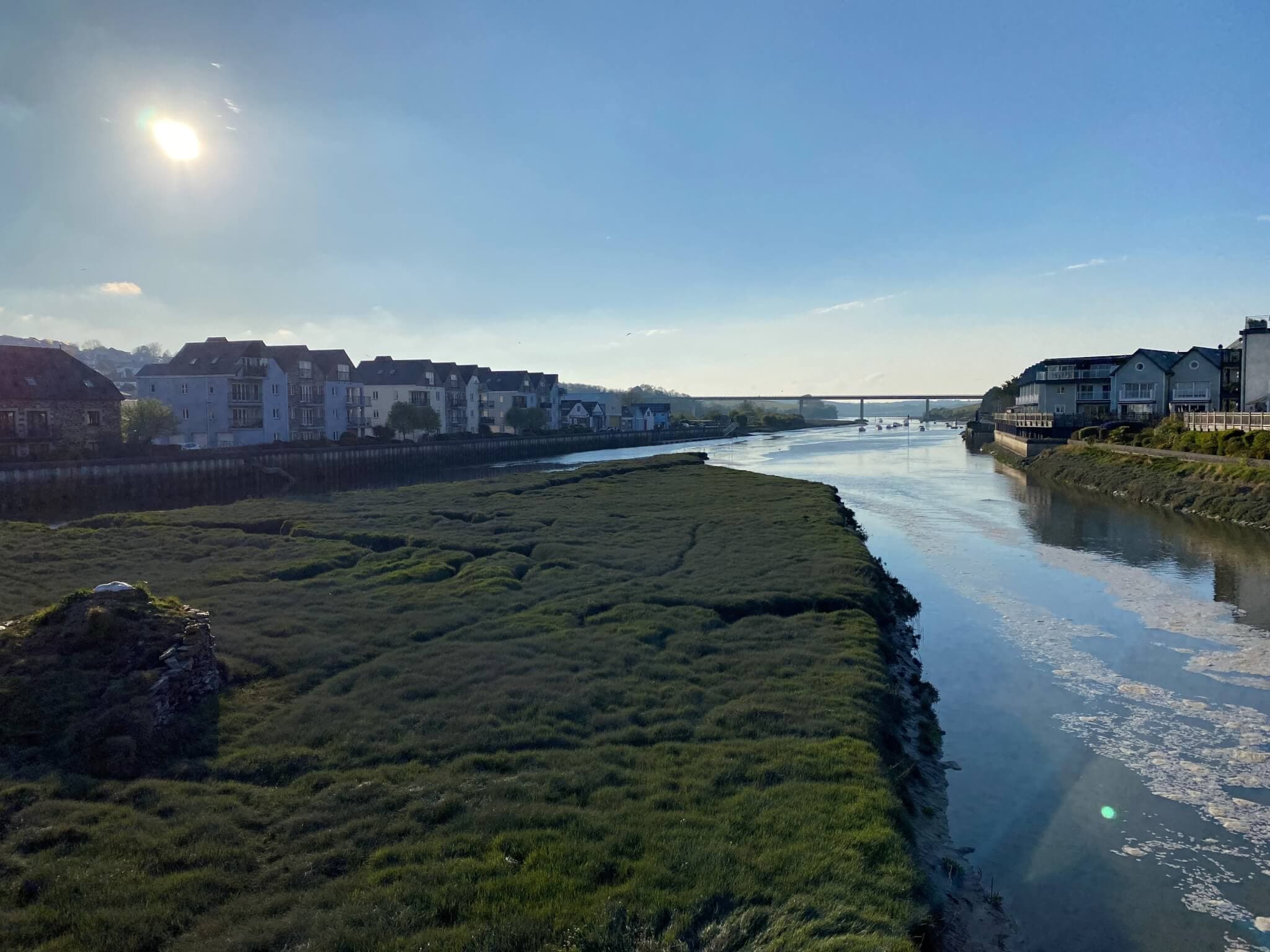 A serene river under a clear sky, flanked by grassy banks and buildings, with a bridge visible in the distance. - Home Instead
