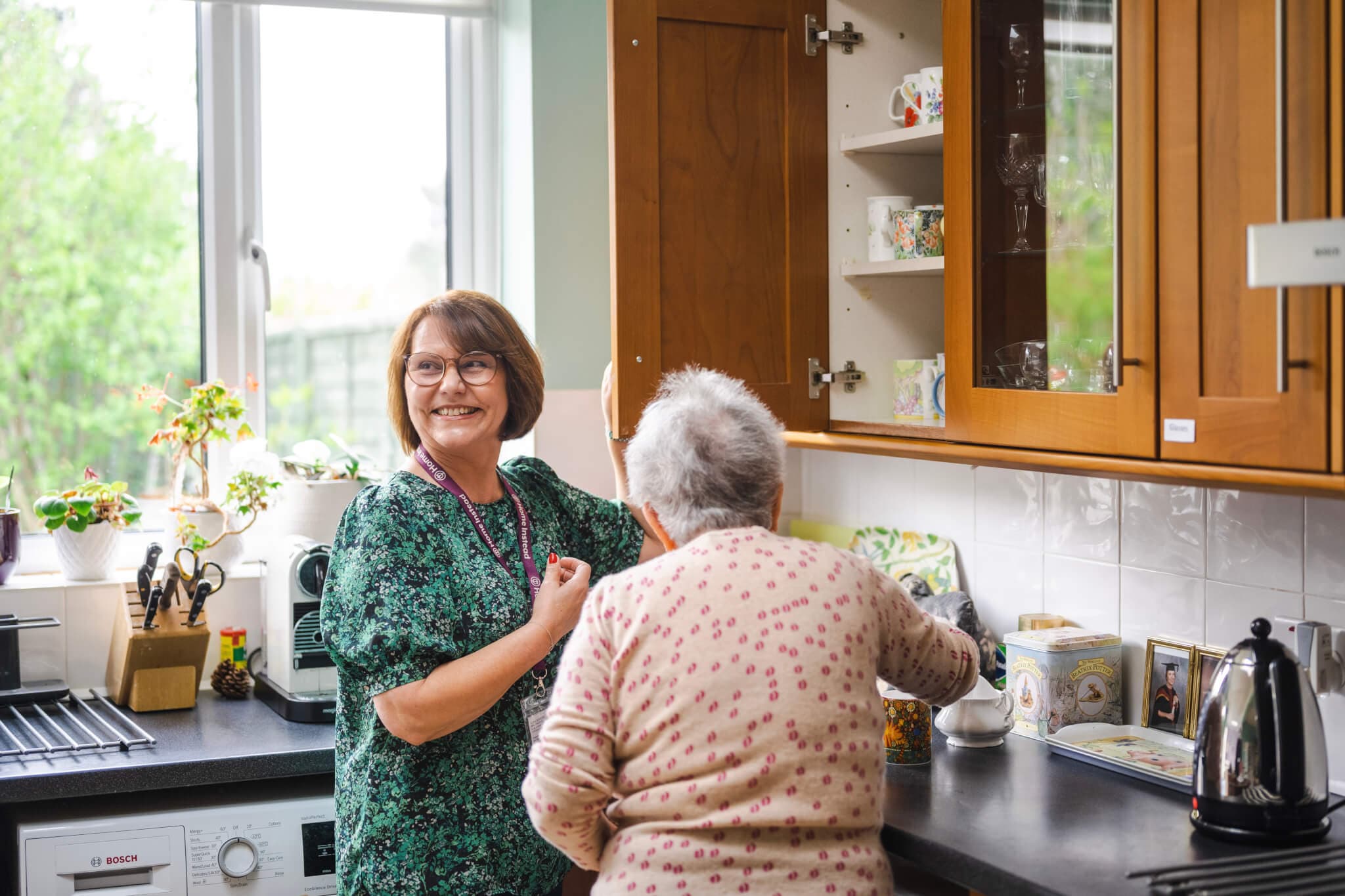 A smiling woman assists an elderly person in a kitchen with wooden cabinets and various items on the countertop. - Home Instead