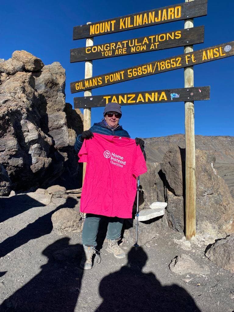 A person holds a pink shirt at the summit of Mount Kilimanjaro in front of signs indicating different peak points. - Home Instead