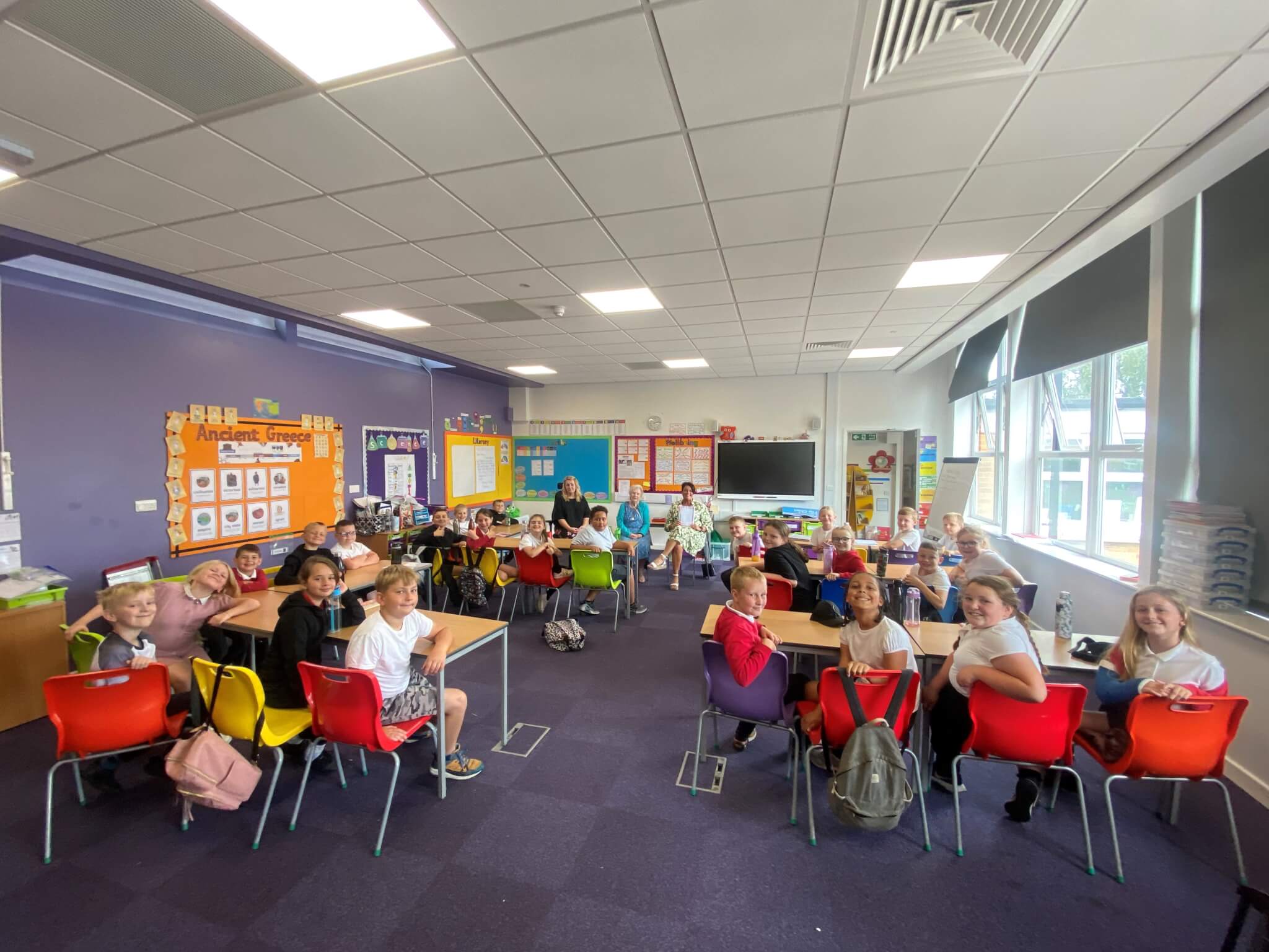A classroom with students and a teacher seated at tables, colorful chairs, and educational posters on the walls. - Home Instead