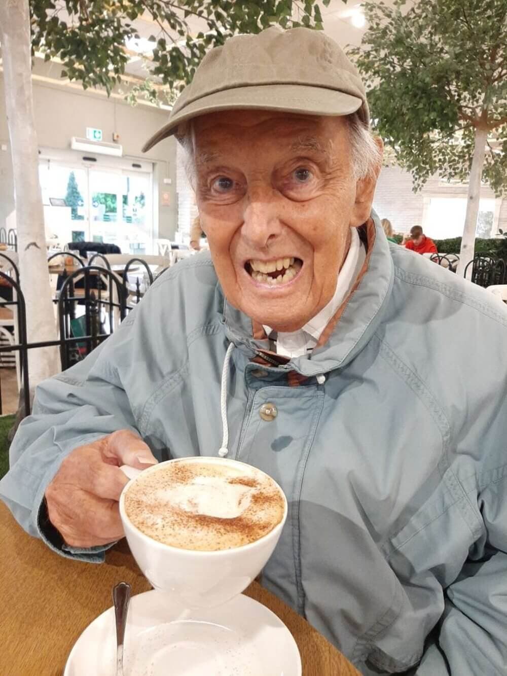 An elderly man in a cap and jacket smiles, holding a cup of coffee with whipped cream, at a cafe. - Home Instead