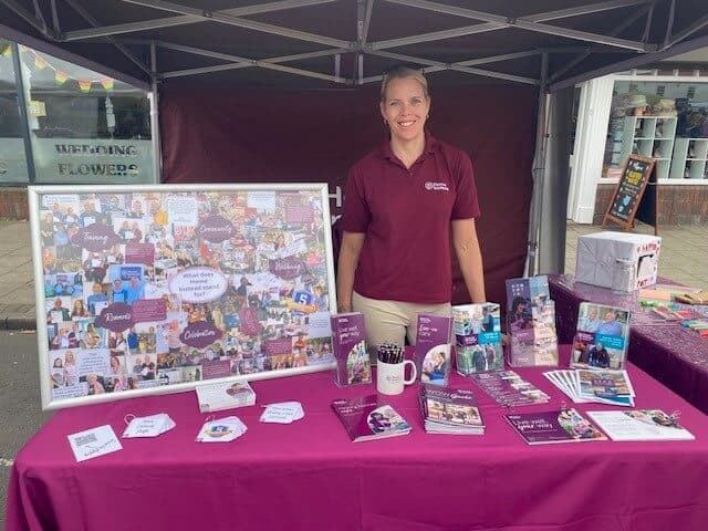 A woman in a maroon shirt stands behind a table with informational materials and a display board at an outdoor booth. - Home Instead