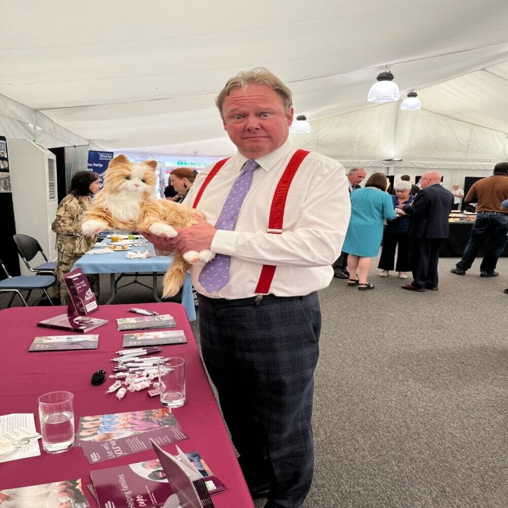 A man with red suspenders holds a robotic cat at a booth in a busy event tent. - Home Instead