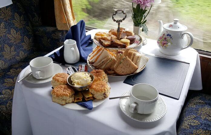 A table with scones, sandwiches, tea, and condiments set for afternoon tea by a train window. - Home Instead