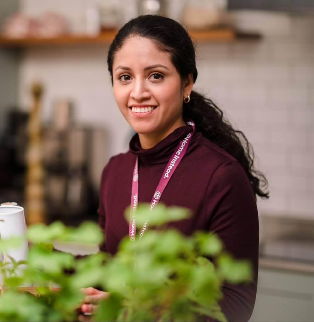A woman with dark hair wearing a maroon top smiles in a kitchen, with plants in the foreground. - Home Instead