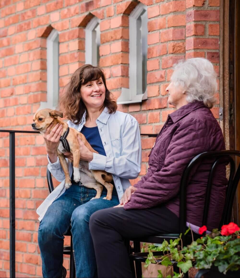 Two women sitting on a porch, one holding a small dog. They are smiling and talking. The background is a brick wall. - Home Instead