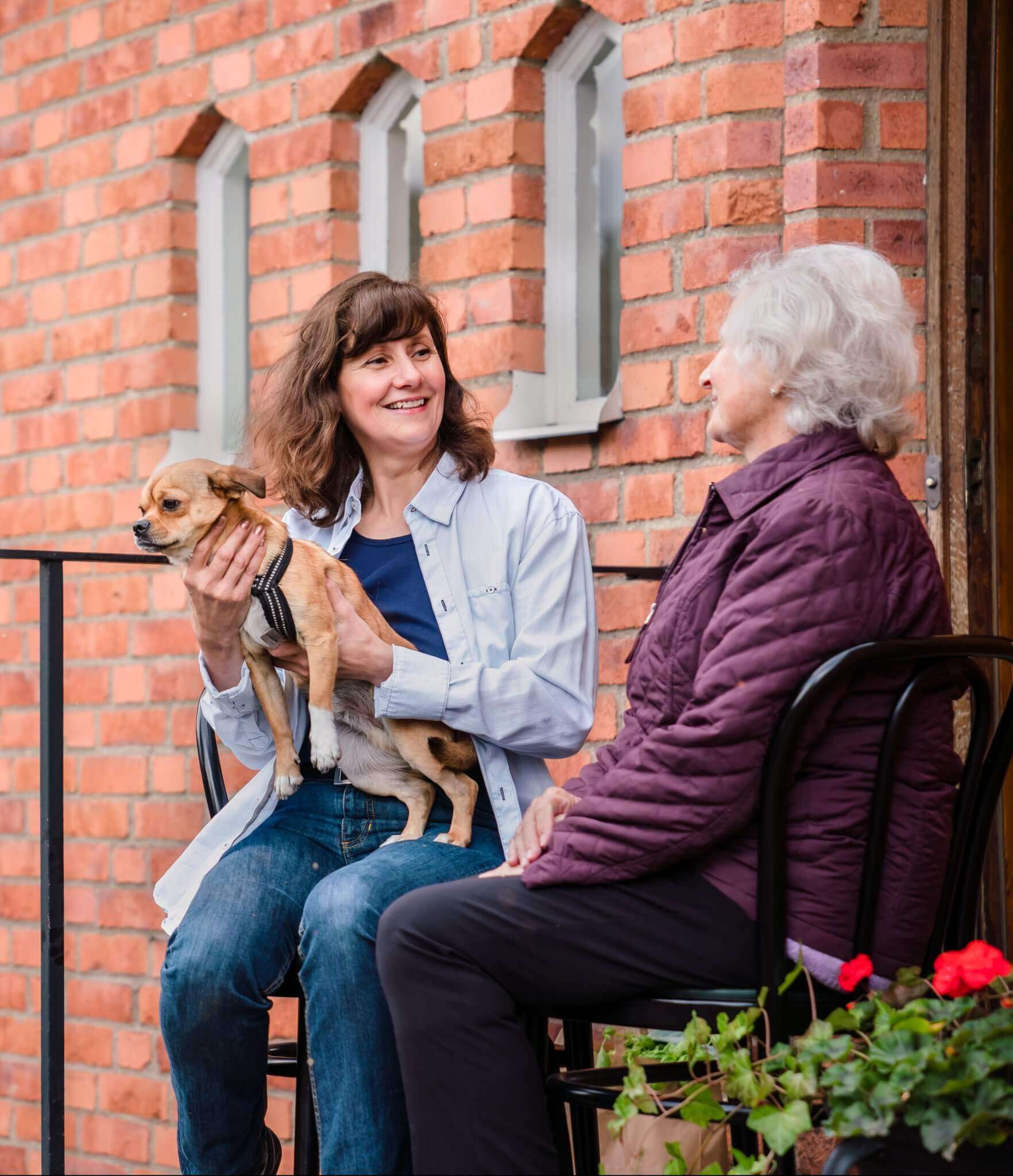Two women sitting on a porch, one holding a small dog. They are smiling and talking. The background is a brick wall. - Home Instead