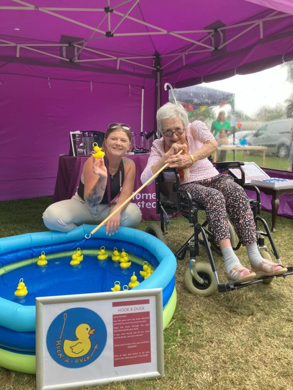 Two people, one elderly in a wheelchair, enjoy a 'Hook a Duck' game in a purple tent at an outdoor event. - Home Instead