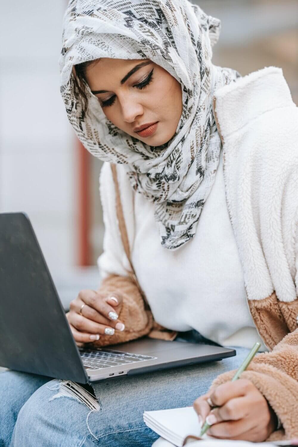 A person wearing a hijab works on a laptop and takes notes on a notepad. - Home Instead