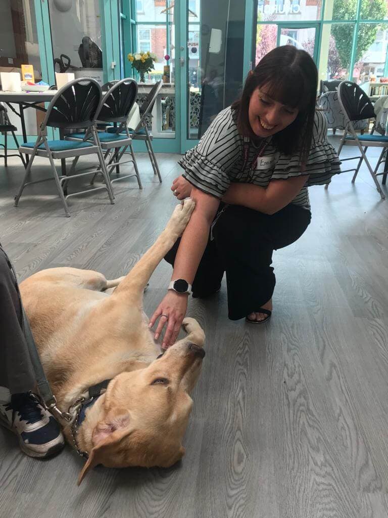 A woman kneels to pet a golden retriever lying on its back indoors. Chairs and tables are visible in the background. - Home Instead