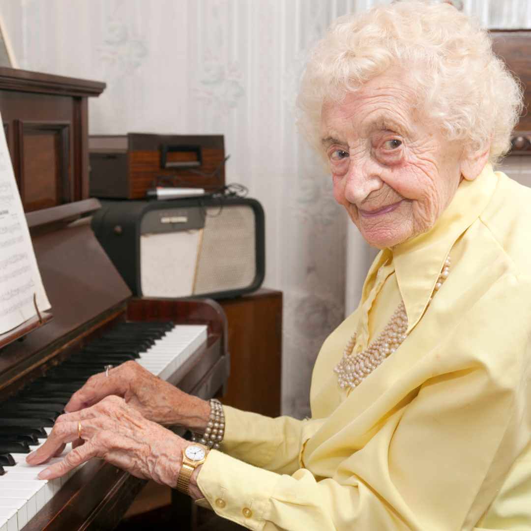 Elderly woman with curly white hair playing the piano, wearing a yellow blouse and smiling warmly. - Home Instead