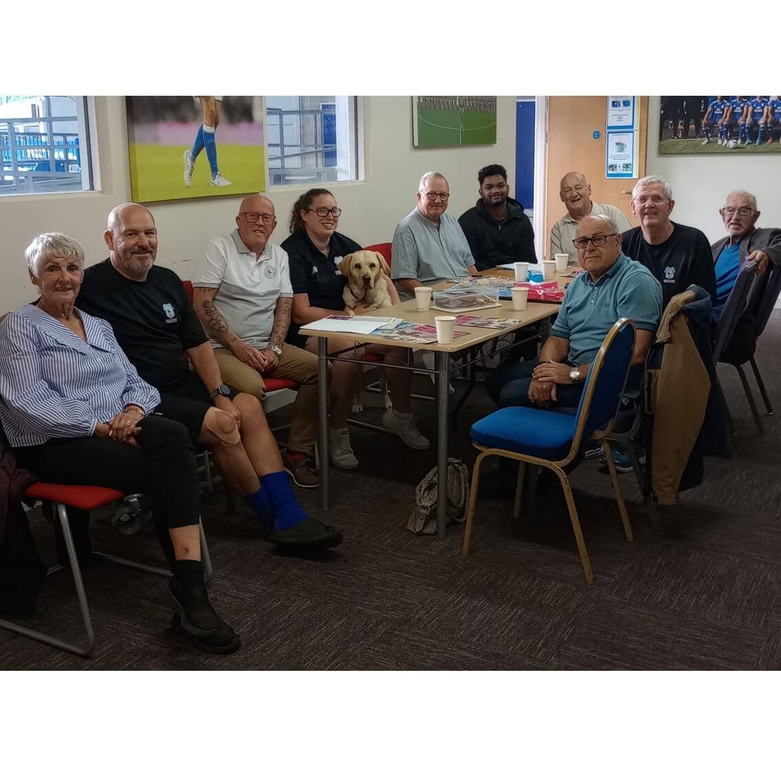 A group of people and a dog sit around a table in a room, smiling at the camera. - Home Instead